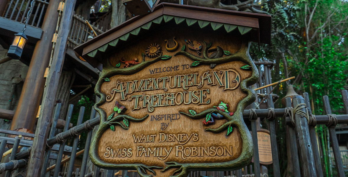 An ornate sign marks the entrance to an attraction. In fancy script, it reads, Welcome to the Adventureland Treehouse inspired by Walt Disney’s Swiss Family Robinson. Around the edges of the sign is a border designed to look like green branches. Above it is the inverted V shape of the eave of an Alpine roof, as if to protect the sign from the elements and also make it look a bit like a house itself.