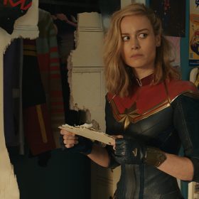 Captain Marvel from The Marvels, played by Brie Larson, is wearing her blue and red suit with a star on the front of it and black gloves with a star on it as she stands in a room in which the walls are covered in posters and drawings. She is holding a piece of white wall in her hands that has papers coming off it as she looks to the side with a questioning look on her face.