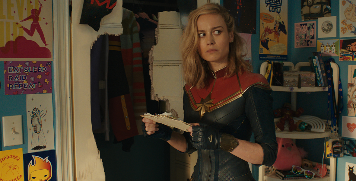 Captain Marvel from The Marvels, played by Brie Larson, is wearing her blue and red suit with a star on the front of it and black gloves with a star on it as she stands in a room in which the walls are covered in posters and drawings. She is holding a piece of white wall in her hands that has papers coming off it as she looks to the side with a questioning look on her face.