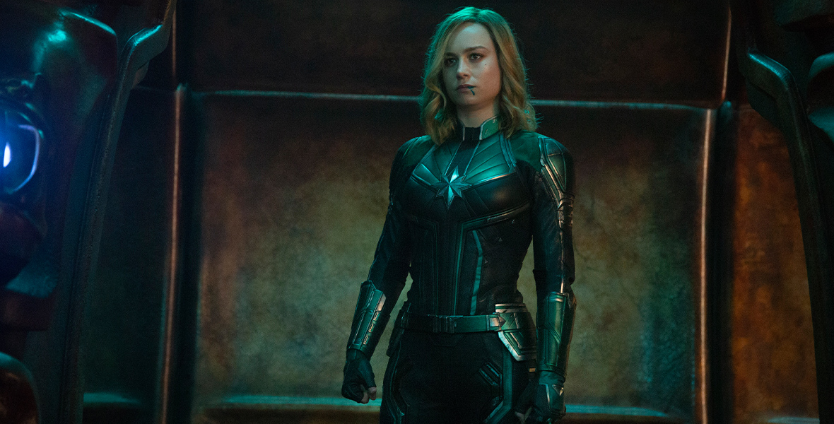 Captain Marvel from Captain Marvel, played by Brie Larson, is standing in front of a metal-looking wall in a black suit with a star on the front and black gloves as blue light shines on her and blood drips from her mouth. 