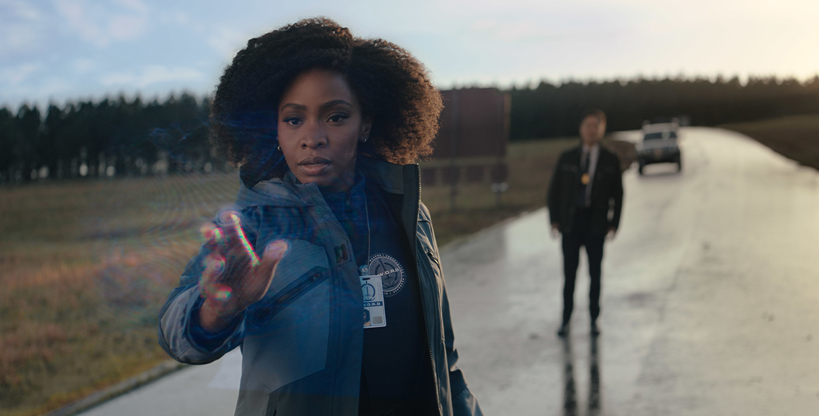 Captain Monica Rambeau from WandaVision, played by Teyonah Parris, is in a gray jacket and is sticking her hand up in front of her, infiltrating rainbow waves. The backdrop features several trees and a patch of grass. Jimmy Woo, played by Randall Park, is standing behind her, out of focus.