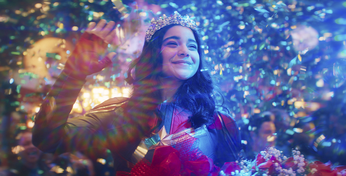 Ms. Marvel, played by Iman Vellani, is dressed in a blue and red suit with a gold star on the front and crown on her head. There is iridescent confetti falling all around her as she holds red and white flowers in her hand.