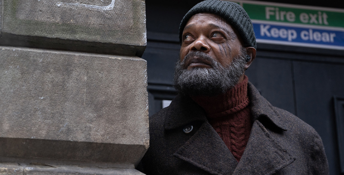 Nick Fury, played by Samuel L. Jackson is dressed in a dark orange turtleneck sweater with a dark-gray coat and hat as he looks to the side and leans on a beige stone wall. A green and blue sign reading “Fire exit Keep clear” is behind him.