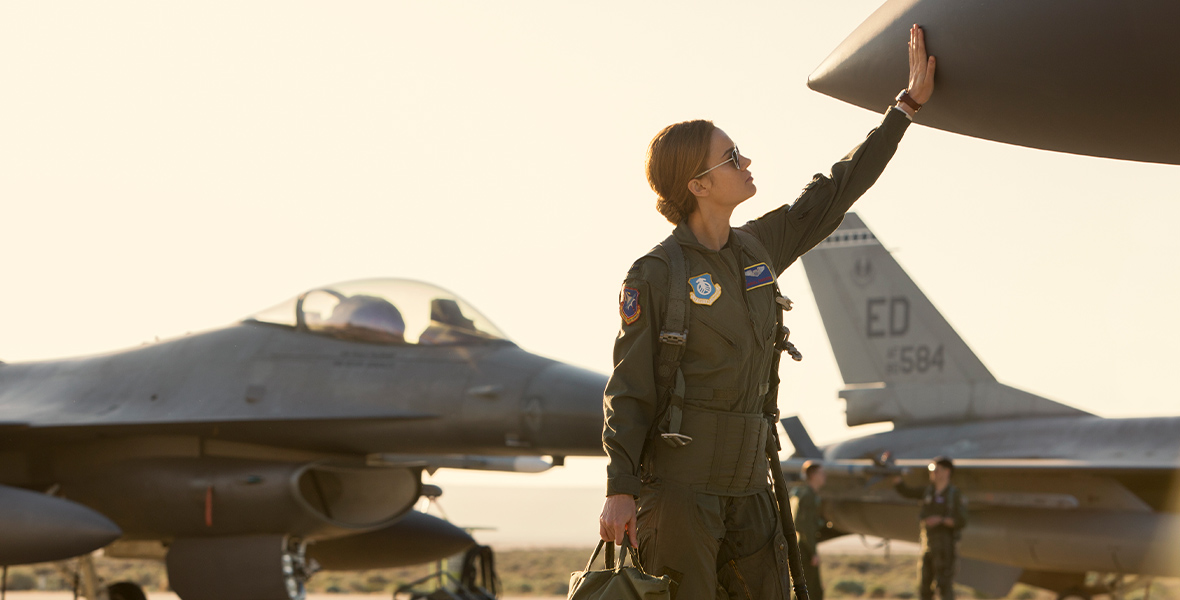 Carol Danvers, played by Brie Larson, in Captain Marvel is wearing a green Air Force uniform while holding a matching bag with patches. She puts one hand up to touch the front of a gray airplane. Two other airplanes are behind her along with two other members of the Air Force in their uniforms.