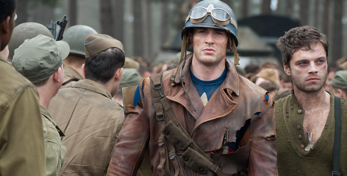 Steve Rogers, played by Chris Evans, in Captain America: The First Avengeris wearing a brown jacket, blue shirt, blue army helmet, and goggles around the helmet. Bucky Barnes, played by Sebastian Stan, stands next to him wearing a green shirt, silver dog tags, and has a cut on his face. Several soldiers surround them.