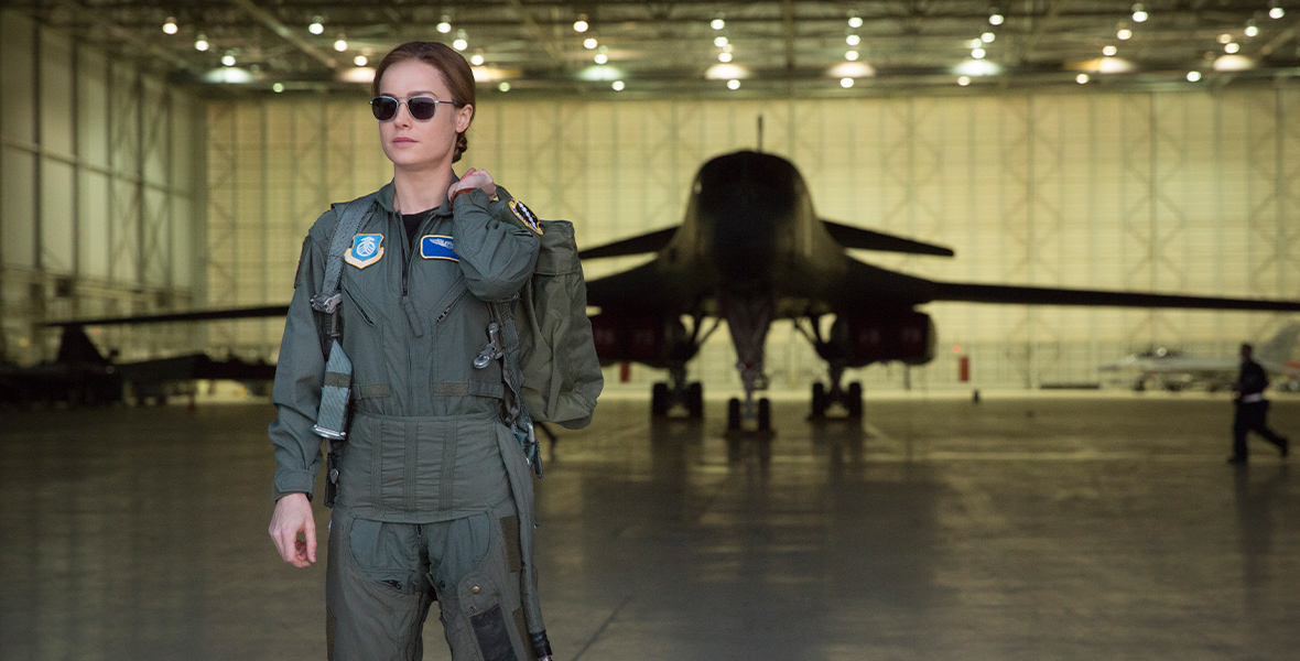 Carol Danvers, played by Brie Larson, in Captain Marvel is wearing a green Air Force uniform with black sunglasses while holding a bag over her shoulder. She is standing in an overhang where a plane is parked behind her.