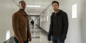 Sam Wilson, played by Anthony Mackie, and Bucky Barnes, played by Sebastian Stan, from The Falcon and The Winter Soldier stand in a long white hallway with a concerned look on their faces. Sam is wearing a light brown jacket with black pants and Bucky is wearing a black jacket, black gloves, and blue jeans.