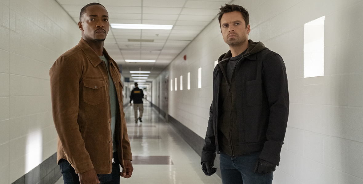 Sam Wilson, played by Anthony Mackie, and Bucky Barnes, played by Sebastian Stan, from The Falcon and The Winter Soldier stand in a long white hallway with a concerned look on their faces. Sam is wearing a light brown jacket with black pants and Bucky is wearing a black jacket, black gloves, and blue jeans.