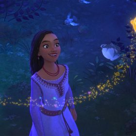 In this still image from Walt Disney Feature Animation’s Wish, Asha stands in a dark forest, smiling up at Star, the round and golden wishing star she has inadvertently conjured from the sky. Star has five small points: two hands, two feet, and a peak above his round face, which includes just two eyes and a mouth. Star is trailed by a scattering of golden stardust. Three birds are also seen around Asha, one in a tree branch and two flying.