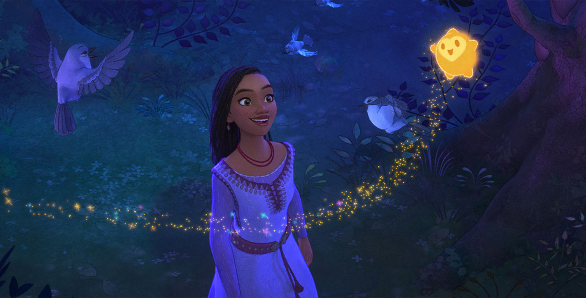 In this still image from Walt Disney Feature Animation’s Wish, Asha stands in a dark forest, smiling up at Star, the round and golden wishing star she has inadvertently conjured from the sky. Star has five small points: two hands, two feet, and a peak above his round face, which includes just two eyes and a mouth. Star is trailed by a scattering of golden stardust. Three birds are also seen around Asha, one in a tree branch and two flying.