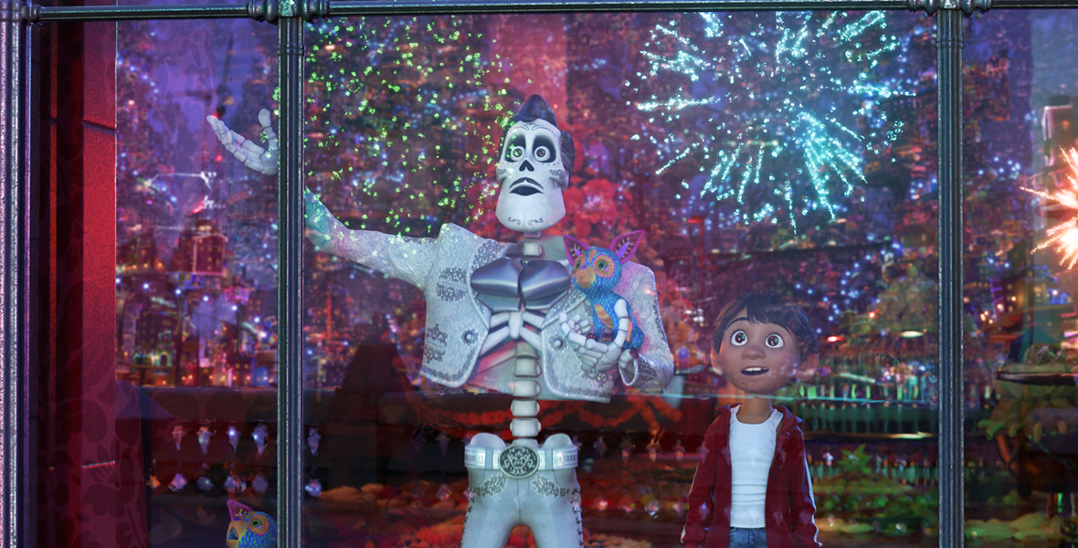 In an image from Disney and Pixar’s Coco, Ernesto de la Cruz (voiced by Benjamin Bratt), left, and Miguel (voiced by Anthony Gonzalez), right, are standing at a window overlooking a huge display of fireworks. Ernesto’s hand is up gesturing at the display, and Miguel’s eyes are full of wonder.