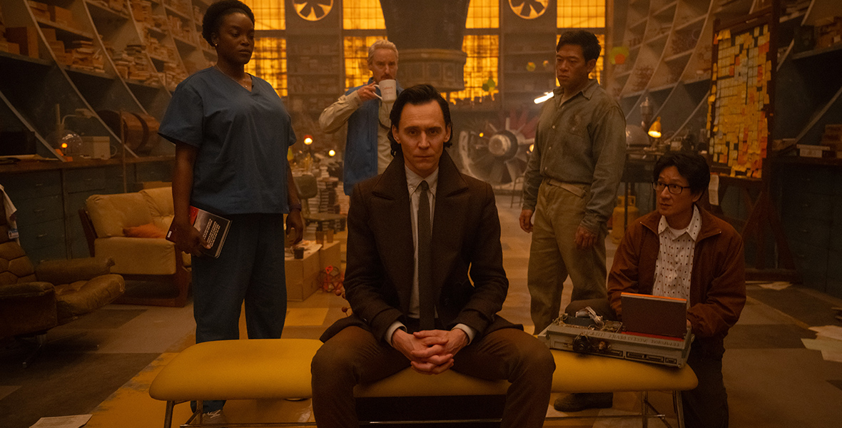 In an image from Marvel Studios’ Loki, from left to right, Hunter B-15 (Wunmi Mosaku), Mobius (Owen Wilson), Loki (Tom Hiddleston), Casey (Eugene Cordero), and O.B. (Ke Huy Quan) are together in a dimly lit room with many bookshelves. Mobius is drinking from a mug; Loki is sitting on a bench; O.B. is typing at a laptop.