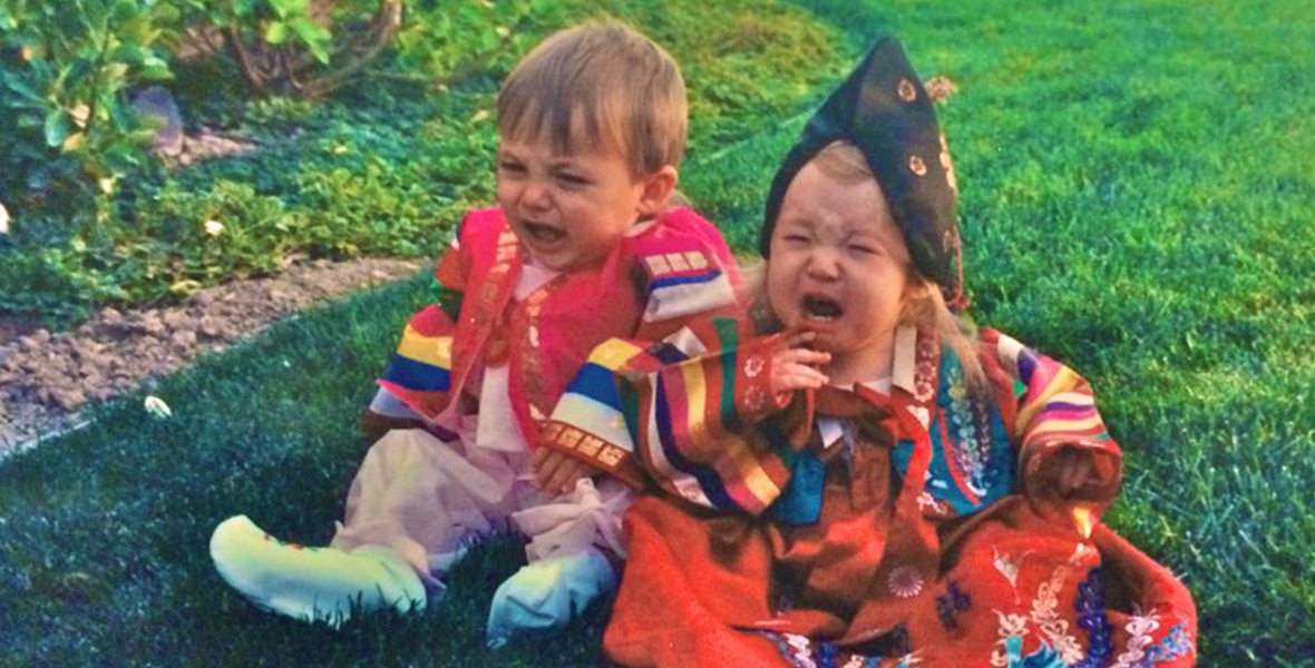 Two toddlers sit on some grass crying. One is wearing a red dress with a black headdress and the other is wearing light pink pants with a hot pink jacket.