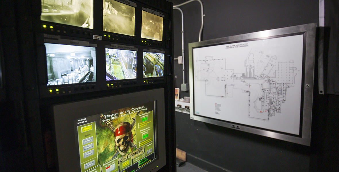 Six small monitors and one larger monitor are mounted on a wall inside the Pirates of the Caribbean control room. Attraction blueprints are mounted on another wall.