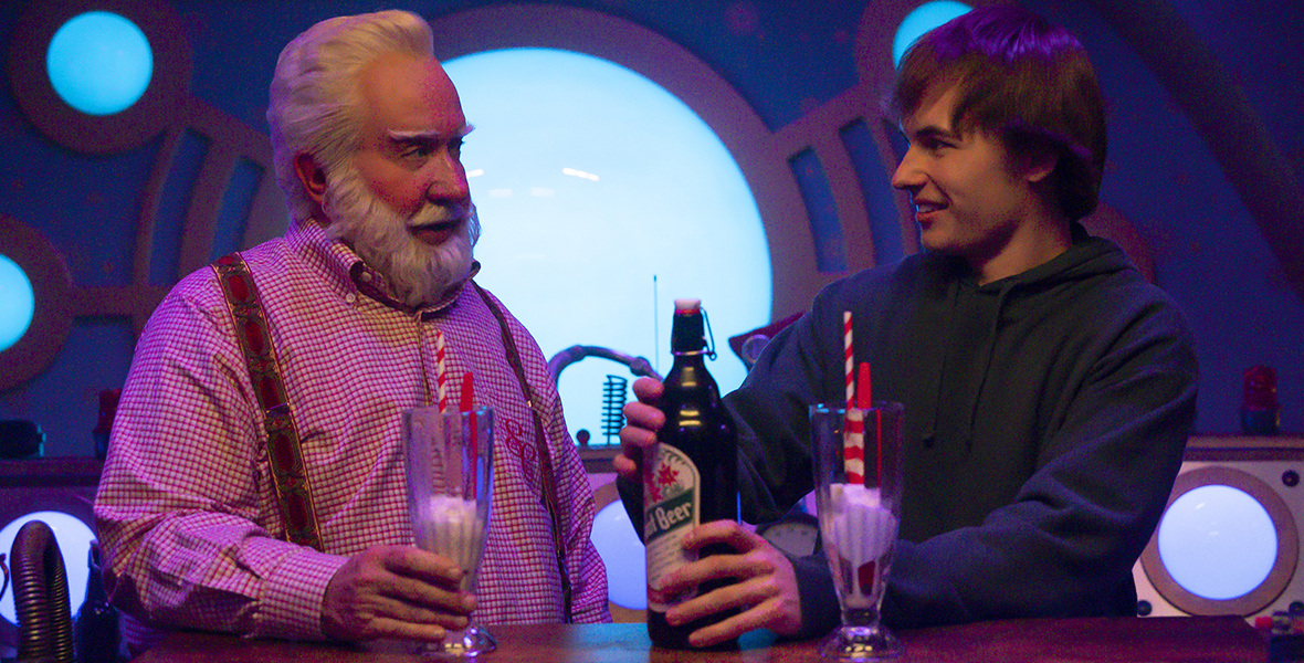 In an image from Disney+’s The Santa Clauses season 2, Scott Calvin, a.k.a. Santa Claus (Disney Legend Tim Allen), left, and his son Cal (Austin Kane), right, are standing at a bar, making themselves root beer floats. Scott is wearing a gingham long-sleeved shirt and suspenders; Cal is wearing a dark hoodie. They’re looking at each other.