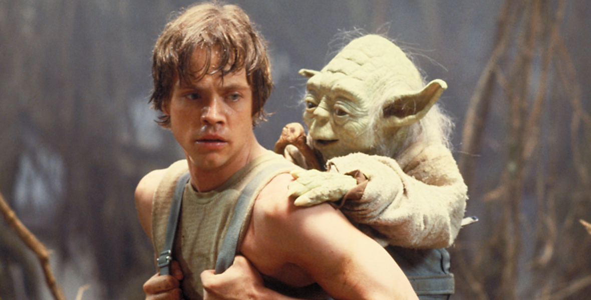 In an image from Lucasfilm’s Star Wars: The Empire Strikes Back, Luke (Disney Legend Mark Hamill) is standing in a swamp, with Jedi Master Yoda (voiced by Frank Oz) strapped to his back. Luke has a determined look on his face.