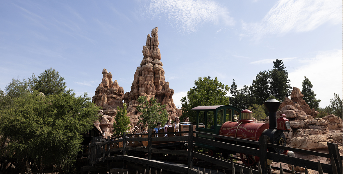 In an image from season 2 of Disney+’s Behind the Attraction, Big Thunder Mountain Railroad is seen, with a ride vehicle full of guests crossing over a bridge of some kind. It’s a bright, sunny day and there are trees and other foliage surrounding this section of the attraction.