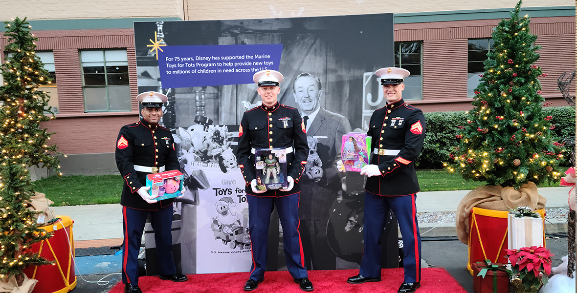 A trio of Marines hold toys and stand on a red carpet. They are posing for a photo in front of a backdrop of Walt Disney and information about The Walt Disney Company's longstanding relationship with the Marine Toys for Tots program.