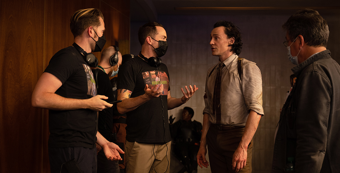 In a behind-the-scenes image from Marvel Studios’ Loki, Tom Hiddleston is seen on the set of the series, surrounded by several producers and other crew members—a few of whom are wearing masks. They’re gesturing and talking, as if between takes during the shoot.