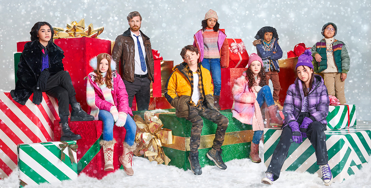 A promo image for Disney Branded Television’s The Naughty Nine featuring Winslow Fegley as Andy, Camila Rodriguez as Dulce, Derek Theler as Bruno, Deric Mccabe as Jon Anthony, Clara Stack as Rose, Anthony Joo as Lewis, Ayden Elijah as Albert, Imogen Cohen as Ha-Yoon, and Madilyn Kellam as Laurel. The group is sitting on or standing near giant wrapped gifts, on a floor covered in snow. They’re all wearing jackets; some wear hats, gloves, and scarves.