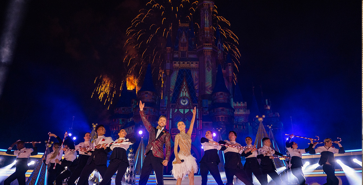 In an image from ABC’s The Wonderful World of Disney: Magical Holiday Celebration, Derek Hough and Julianne Hough are seen performing on stage in front of Cinderella Castle at Magic Kingdom at Walt Disney World Resort. Dancers flank them on either side, while fireworks glow in the skies above them.