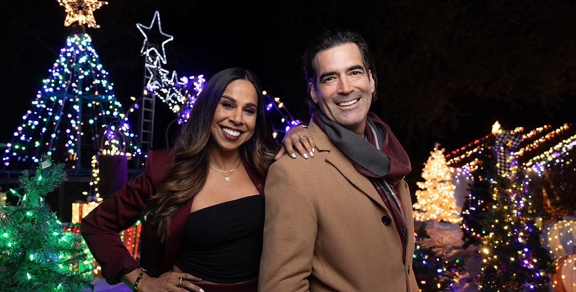 In an image from ABC’s The Great Christmas Light Fight, hosts and judges Taniya Nayak and Carter Oosterhouse stand in front of a display from an episode featured this season. Plentiful twinkling holiday lights can be seen behind Nayak and Oosterhouse; she is wearing a maroon suit, while he is wearing a tan suit jacket and maroon and grey scarf.