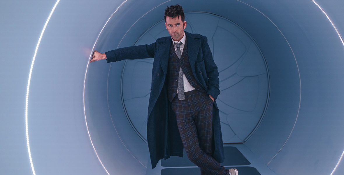 In an image from the 60th anniversary specials for BBC’s Doctor Who on Disney+, the Fourteenth Doctor (David Tennant) is leaning against one of the hallways in the TARDIS, with a serious look on his face. He’s wearing his signature trench coat, tie, and Chuck Taylor sneakers.