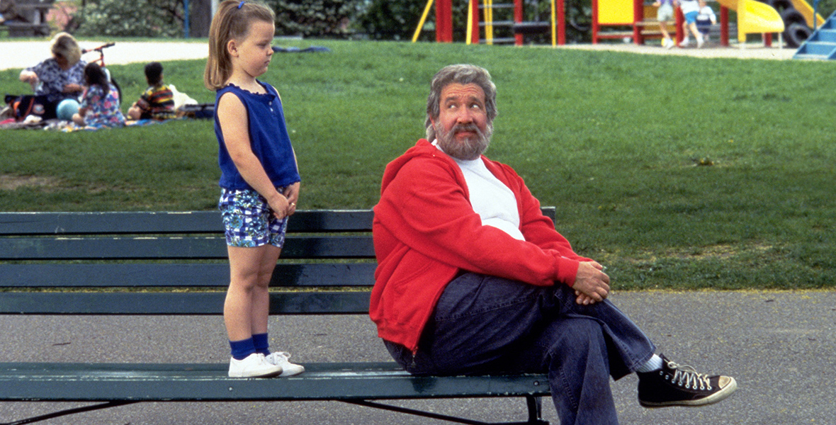 In an image from 1994’s The Santa Clause, a little girl stands on a park bench staring at Scott Calvin (Disney Legend Tim Allen), who has the unmistakable appearance of Santa Clause. Scott—who, for his part, is trying to ignore the little girl—is wearing a red hooded sweatshirt and blue pants.