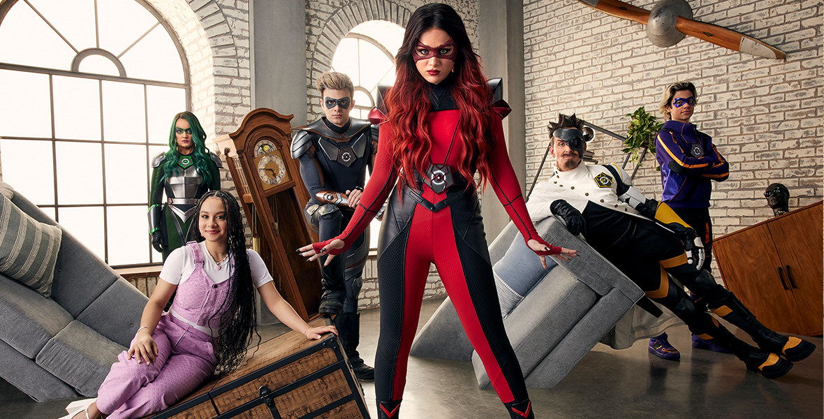 A promotional image for Disney Channel’s The Villains of Valley View features Isabella Pappas as Havoc/Amy, Lucy Davis as Eva/Surge, James Patrick Stuart as Vic/Kraniac, Malachi Barton as Colby/Flashform, Reed Horstmann as Jake/Chaos, and Kayden Muller-Janssen as Hartley. They are standing on a set that features furniture coming out of the floor at odd angles, as if from another dimension. Everyone except Muller-Janssen is wearing typical “villains”-style outfits—colorful spandex suits, masks, and the like.