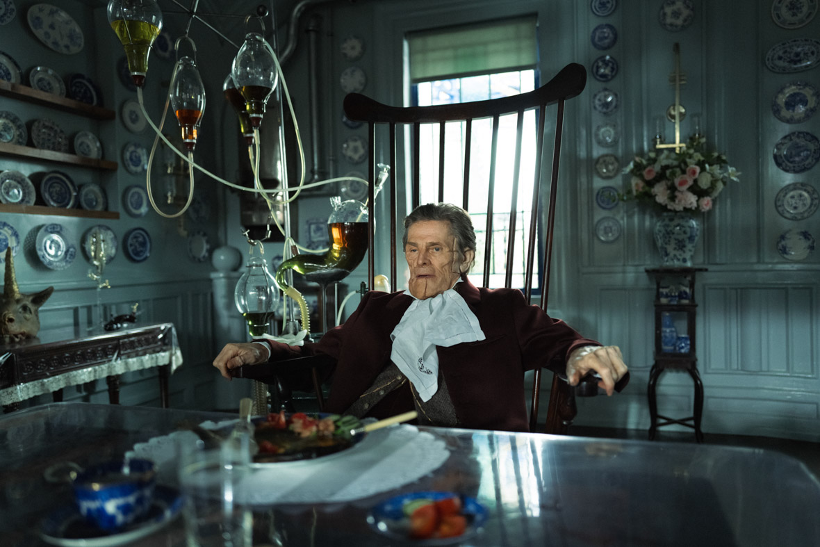 Willem Dafoe plays Dr. Godwin Baxter in Poor Things. His face is disfigured and he sits in an oversized chair. Next to him are several glass orbs and tubes that produce gas.
