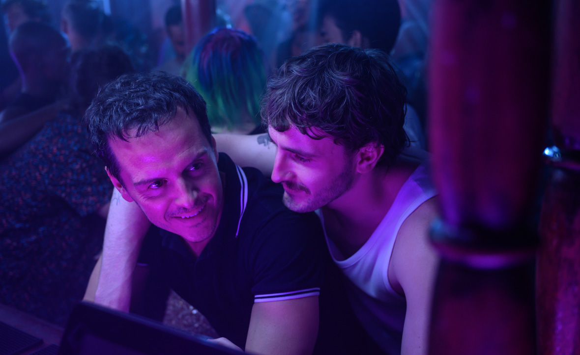 In a scene from All of Us Strangers, Adam, played by Andrew Scott, leans forward in a crowded bar. Harry, played by Paul Mescal, drapes his right arm over Adam's shoulder.
