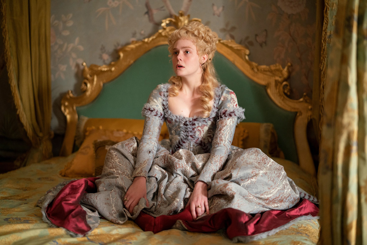 Catherine, played by Elle Fanning, sits cross-legged on her bed in a scene from The Great. She is wearing an ornate brocade dress and appears confused or exasperated.