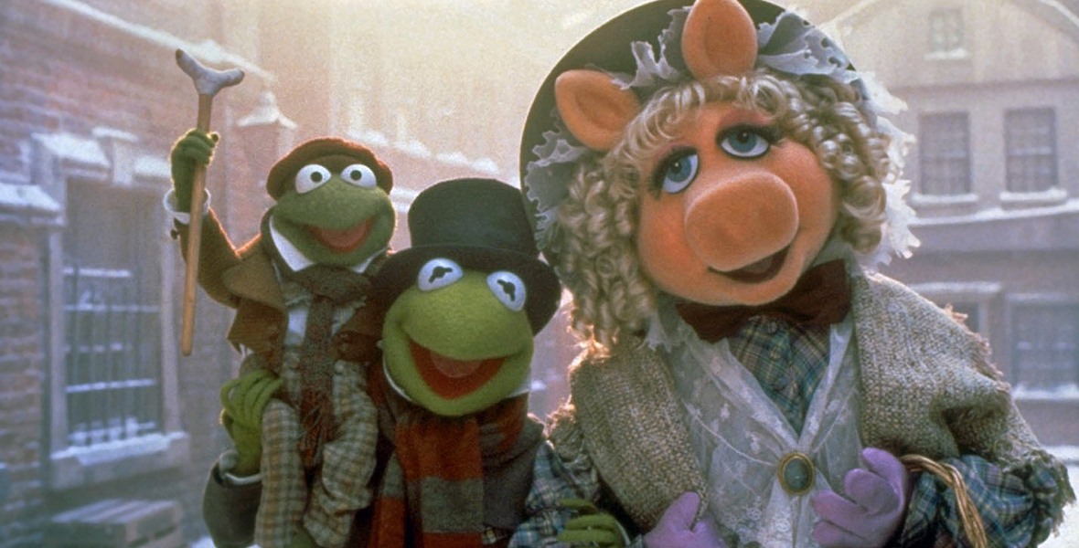 In an image from The Muppet Christmas Carol, Tiny Tim (Robin the frog) is sitting atop the shoulder of his dad, Bob Cratchit (Kermit the Frog), holding his crutch aloft; they’re standing next to Emily Cratchit (Miss Piggy); everyone wears 19th century clothing and they’re standing on a snowy London street.