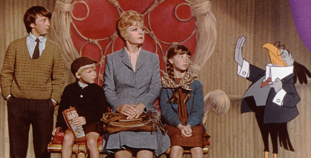 In an image from Disney’s Bedknobs and Broomsticks, Eglantine Price (Disney Legend Angela Lansbury) is sitting on an animated throne surrounded by the children she’s been charged with looking after during the London blitz (played by Ian Weighill, Roy Snart, and Cindy O’Callaghan). To the right is a bird dressed in a suit and glasses; he’s gesturing seriously with his wing.
