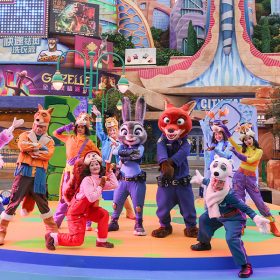 During the opening ceremony for Zootopia at Shanghai Disney Resort, Judy Hopps and Nick Wilde stand in the center of a stage, with a variety of performers posing around them. Each performer wears a hat designed to look like an animal head, with a coordinating outfit. Behind the stage is Zooptopia’s City Hall, along with various skyscraper-like buildings, making up the city’s skyline.