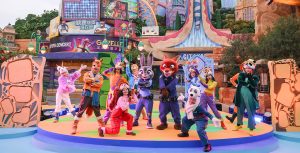 During the opening ceremony for Zootopia at Shanghai Disney Resort, Judy Hopps and Nick Wilde stand in the center of a stage, with a variety of performers posing around them. Each performer wears a hat designed to look like an animal head, with a coordinating outfit. Behind the stage is Zooptopia’s City Hall, along with various skyscraper-like buildings, making up the city’s skyline.