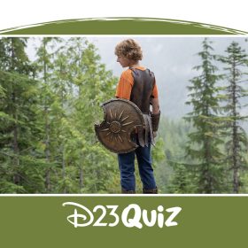 Percy Jackson stands with his back to the viewer, slightly looking over his left shoulder. He is holding a bronze shield in his left hand. In the distance is a thick forest of pine trees