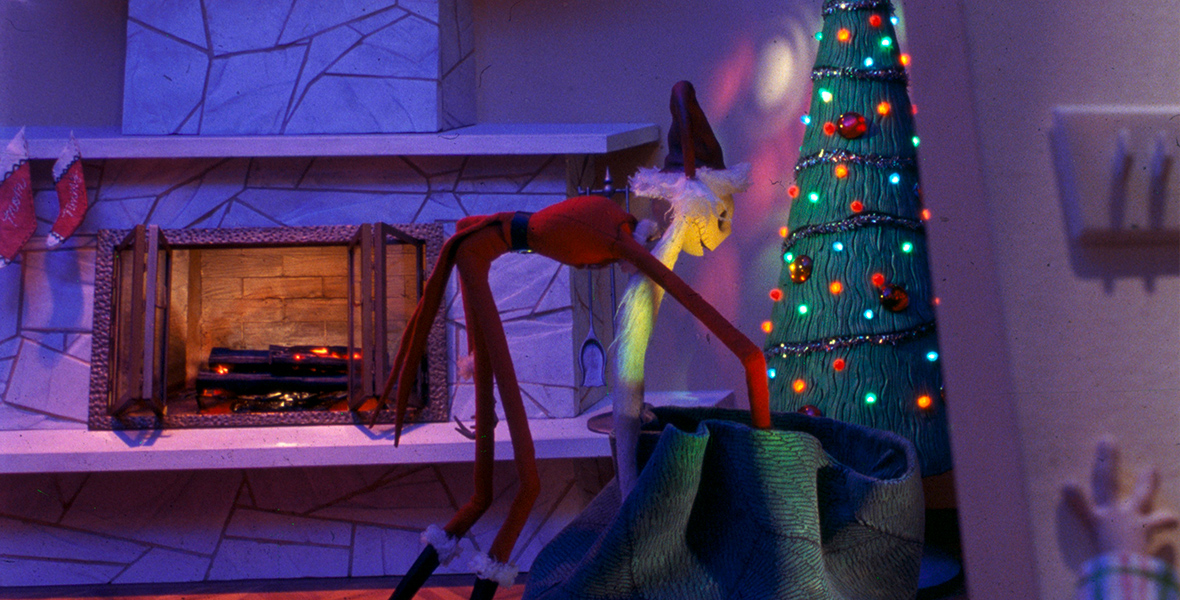 In an image from Tim Burton’s The Nightmare Before Christmas, Jack Skellington (voiced by Chris Sarandon) is dressed like Santa Claus and placing gifts under a Christmas tree. There is a fireplace to his left, above which are hung several stockings.