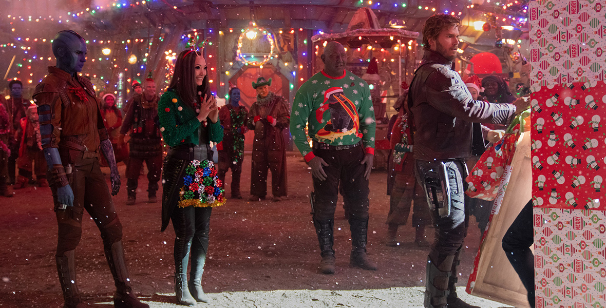 In an image from the Marvel Studios’ Special Presentation The Guardians of the Galaxy Holiday Special, from left to right, Karen Gillan as Nebula, Pom Klementieff as Mantis, Dave Bautista as Drax, and Chris Pratt as Peter Quill/Star-Lord are standing in the middle of Knowhere as Peter begins unwrapping a huge gift. Mantis and Drax are wearing ugly Christmas sweaters, and Christmas lights are strewn every which way.