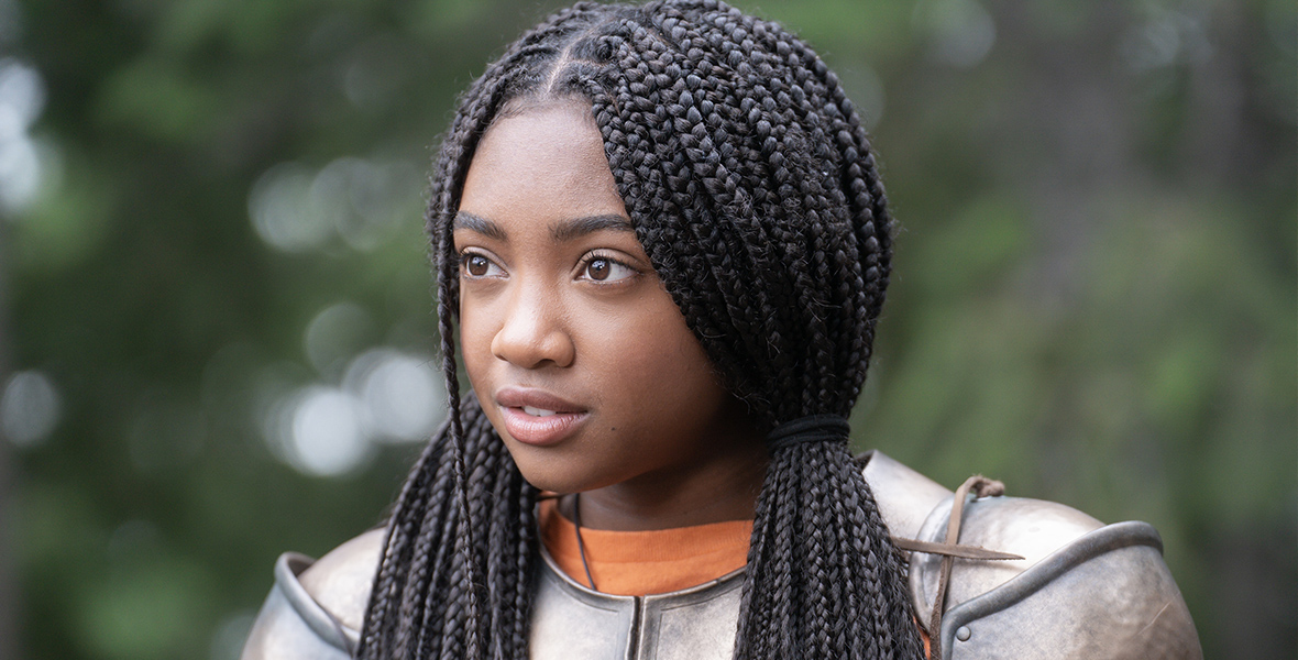 Annabeth Chase (Leah Sava Jeffries) turns and looks toward the left, in a green forest. Her long dark hair is in tight all-over braids and tied back on each side creating loose ponytails. She wears a suit of armor over her orange camp shirt.