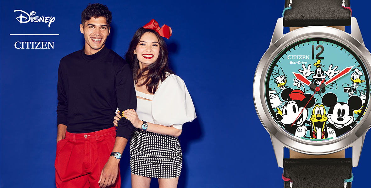 A young man wearing a black turtleneck and red pants stands with a woman with a red bow in her hair, white shirt, and gingham skirt---both are smiling. On the top left is the logo “Disney |Citizen” and to the far right is a large Citizen watch with classic Disney characters on the face.