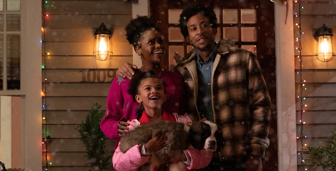Characters from Dashing Through the Snow, including Allison, played by Teyonah Parris; Charlotte, played by Madison Skye Validum; and Eddie, played by Chris “Ludacris” Bridges; stand on the porch of a house that has Christmas lights on it while it snows around them. Charlotte has a Saint Bernard puppy in her arms.