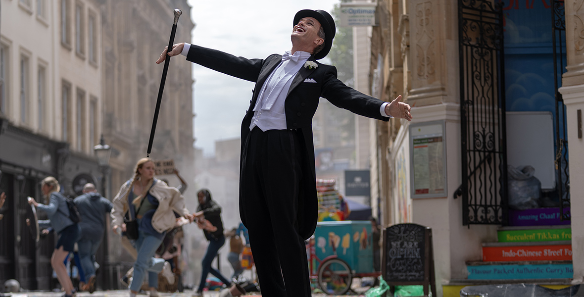 In an image from The Giggle, the third and final special for Doctor Who’s 60th anniversary, the Toymaker (Neil Patrick Harris)—wearing a top hat and tails and carrying a cane—stands in the middle of a busy street, throwing his head back and laughing. Worried people are dashing by him, ostensibly to take cover, as there is debris surrounding the Toymaker on the street.