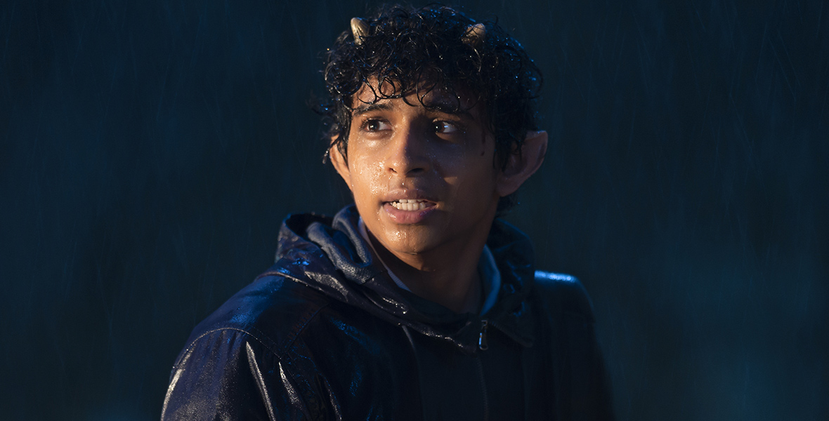 Grover Underwood (Aryan Simhadri) stands looking off at something at left in a darkened rainy wood. His satyr horns and hair are soaked as is his dark hooded jacket.