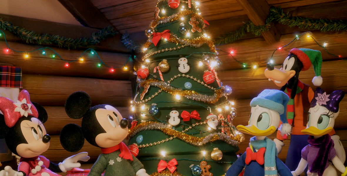 In an image from Mickey Saves Christmas, from left to right, Minnie Mouse, Mickey Mouse, Donald Duck, Goofy, and Daisy Duck surround a Christmas tree inside a log cabin. They all wear holiday finery, and Christmas lights adorn both the tree and the wall behind them.