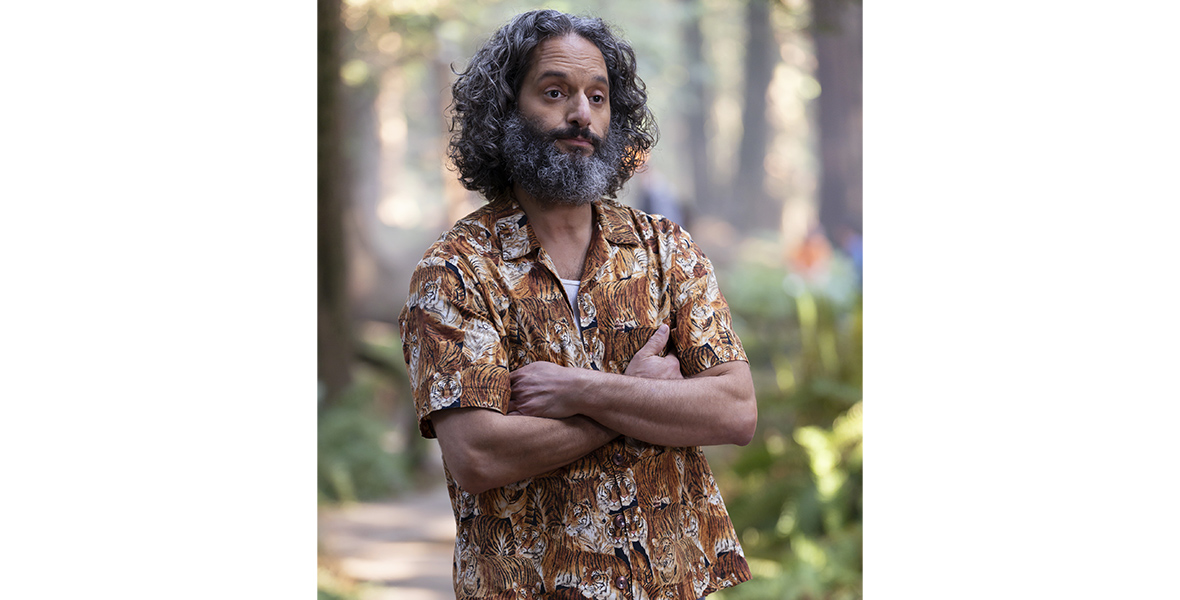 Dionysus aka Mr. D (Jason Mantzoukas) as his alter-ego running Camp Half-Blood stands with his arms crossed and eyebrows raised in seeming judgement. He wears a tiger-printed short-sleeve shirt and jeans with woods visible behind him.