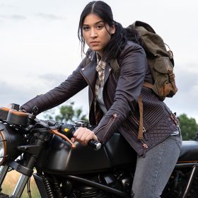 Maya Lopez, played by Alaqua Cox, in Echo sits on a black motorcycle. She’s wearing gray jeans, a blue leather jacket, and a gray backpack and staring toward the camera. In the background is a field and green trees.