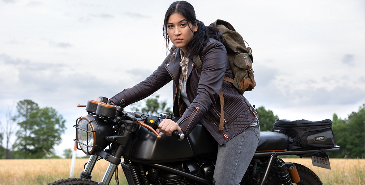 Maya Lopez, played by Alaqua Cox, in Echo sits on a black motorcycle. She’s wearing gray jeans, a blue leather jacket, and a gray backpack and staring toward the camera. In the background is a field and green trees.