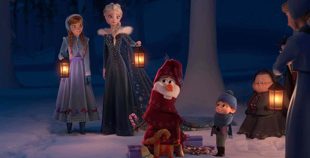 In an image from Walt Disney Animation Studios Olaf’s Frozen Adventure, Anna (voiced by Disney Legend Kristen Bell), left, and Elsa (voiced by Disney Legend Idina Menzel), right, watch as Olaf (Disney Legend Josh Gad) is wrapped up mummy-style in a long red scarf by a small child, standing to his right. The sister pair are each wearing their winter coats and are holding lanterns. There are a few gifts at Olaf’s feet, as if he’s a stand-in for a Christmas tree. Several other people stand off to the right.