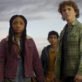 (Left to right) Annabeth Chase (Leah Sava Jeffries) stands looking out at something in the distance. She is wearing a purple corduroy jacked over blue button-up shirt and olive pants, her braided hair pulled partially back. At center in slightly behind is Grover Underwood (Aryan Simhadri), who also looks out in the distance, wearing a dark jacket over mustard yellow shirt, his satyr ears visible atop his brown-haired head. Percy Jackson (Walker Scobell) stands at right, half-way turned toward Annabeth. He wears a green flannel-print shirt and dark pants. All look concerned.